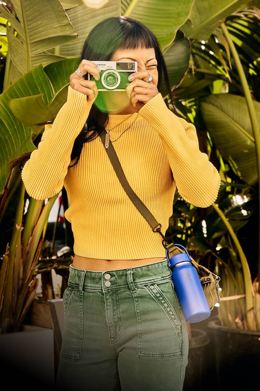 woman holds up camera while outdoors and wears micro hydro mini bottle with a strap