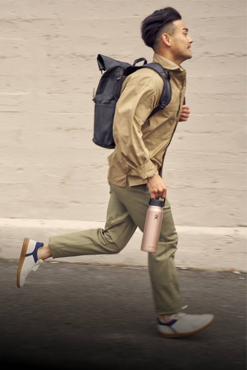 man runs by building wearing backpack and holding hydro flask lightweight water bottle